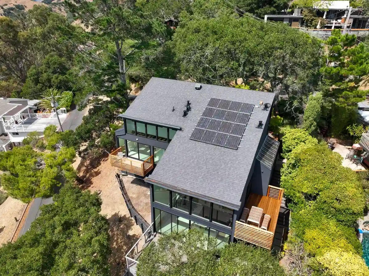 Aerial view of a hillside home surrounded by mature trees, with a dark asphalt shingle roof, rooftop solar panels, and a steep, sloped lot.