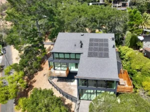 Aerial view of a hillside home with a dark asphalt shingle roof, solar panels, and wood decks surrounded by trees.