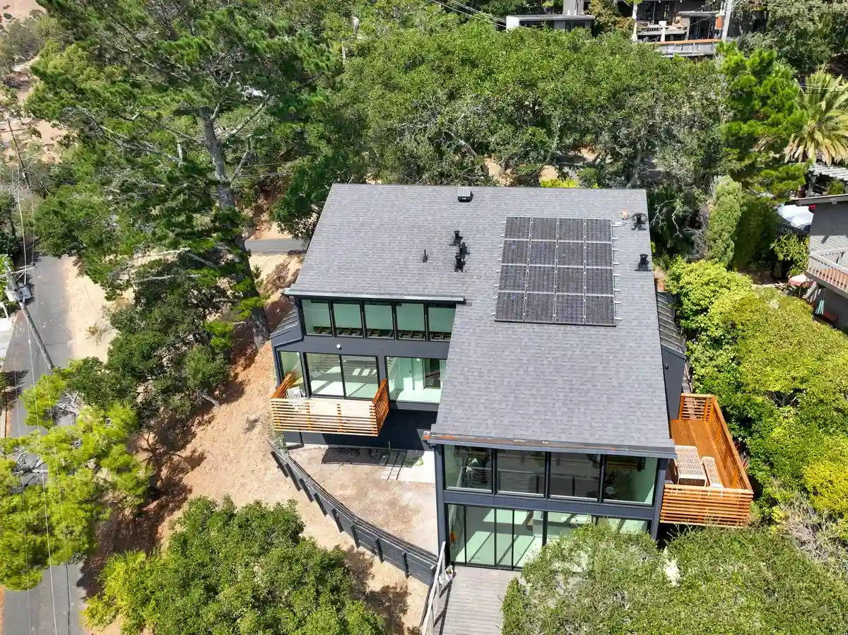 Aerial view of a hillside home with a dark asphalt shingle roof, solar panels, and wood decks surrounded by trees.