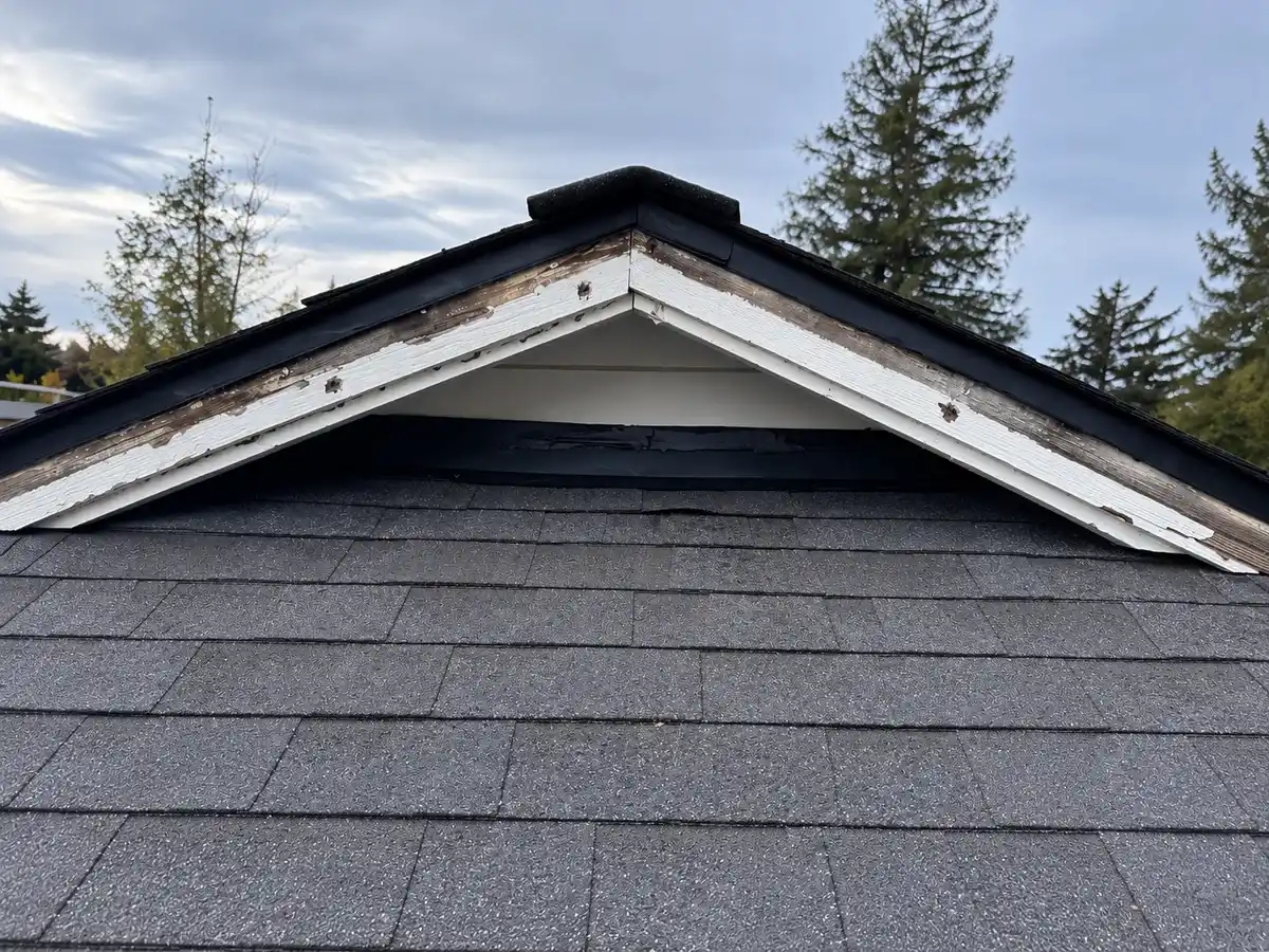Weathered gable trim with peeling paint and exposed wood above asphalt shingles, showing age-related roof wear.