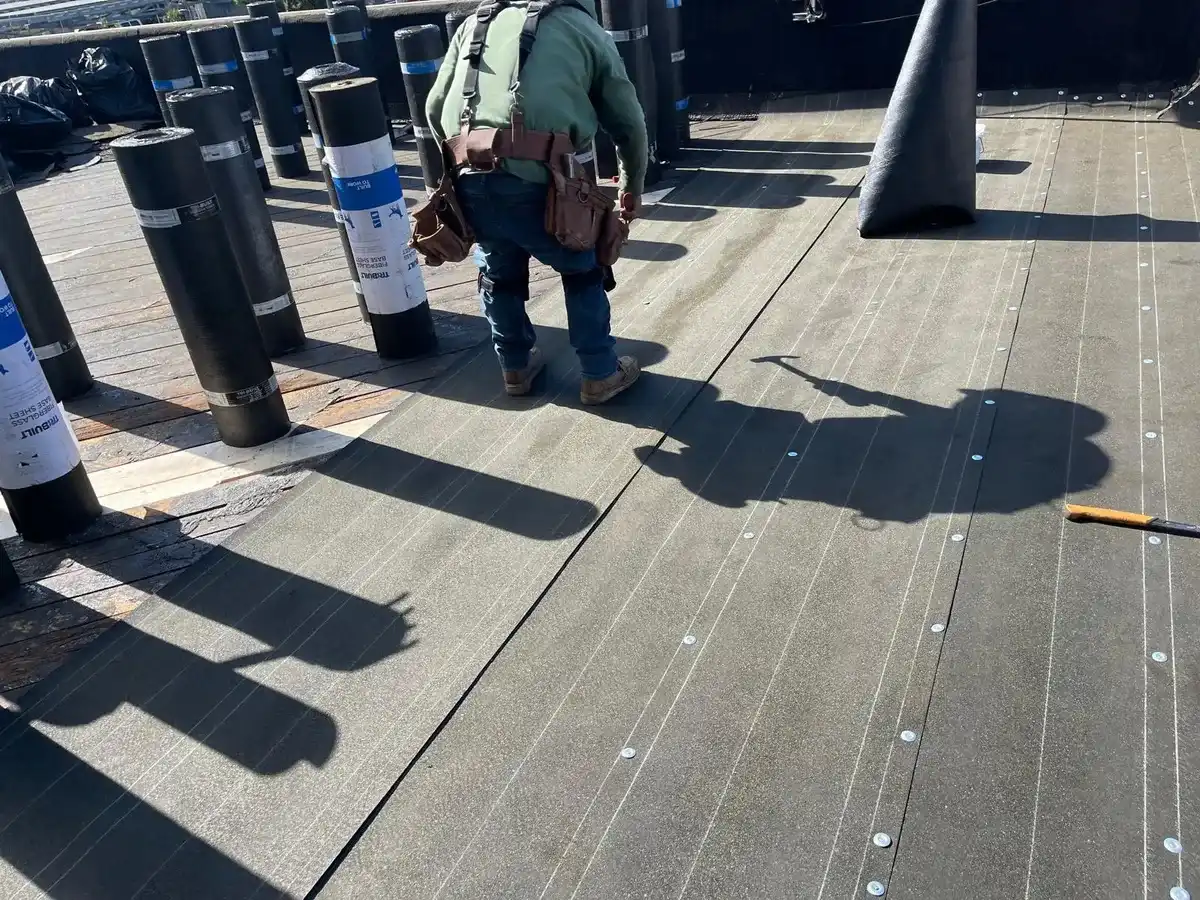 Roofer walking across a flat roof covered in newly fastened base sheet, with rolls of roofing membrane standing upright along the edge.