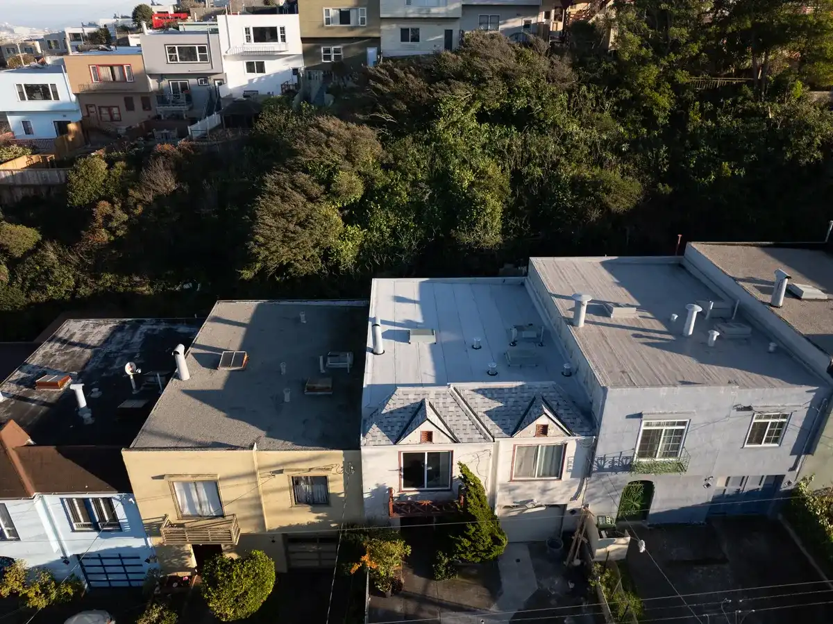 Aerial view of attached hillside homes with flat roofs, roof vents, and a small shingled front gable, backed by dense trees.